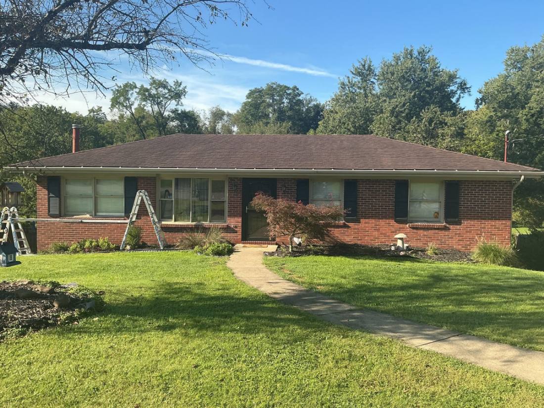 One-story brick home with wide front windows, concrete walkway, manicured lawn, and surrounding trees
