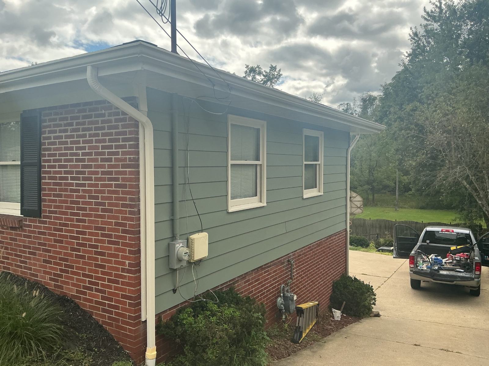Small house with brick foundation and gray siding featuring two windows and driveway