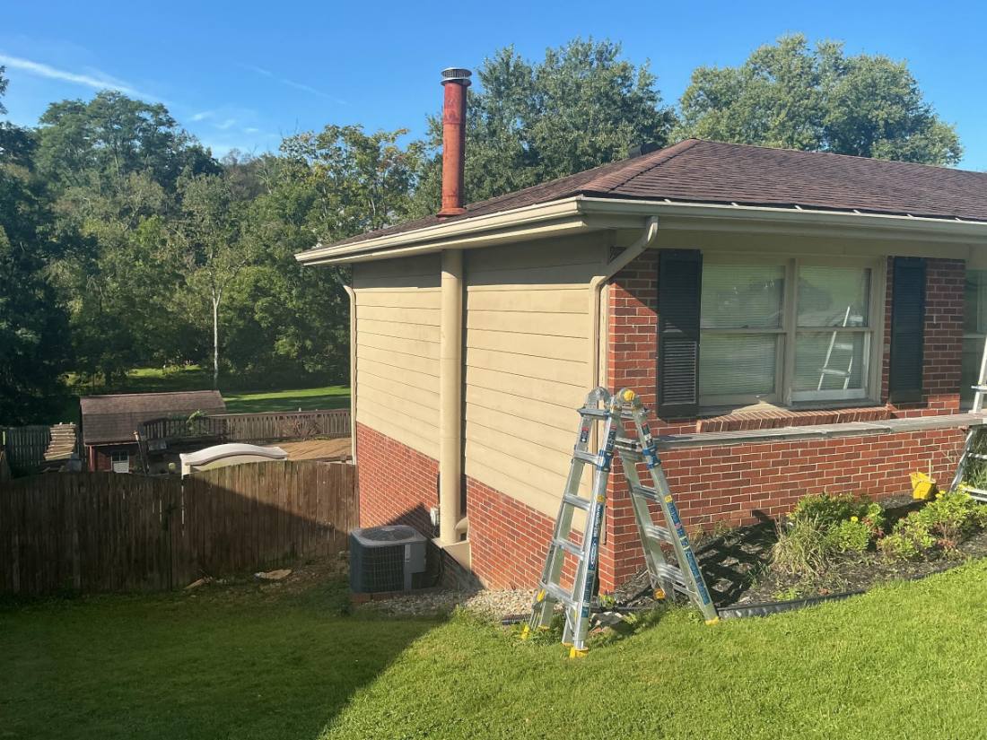 Brick house with cream siding addition, red chimney, ladder leaning against wall, green lawn and trees