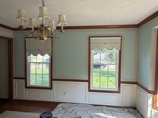 Empty room with light blue walls, wood trim, two windows, brass chandelier, and hardwood floors
